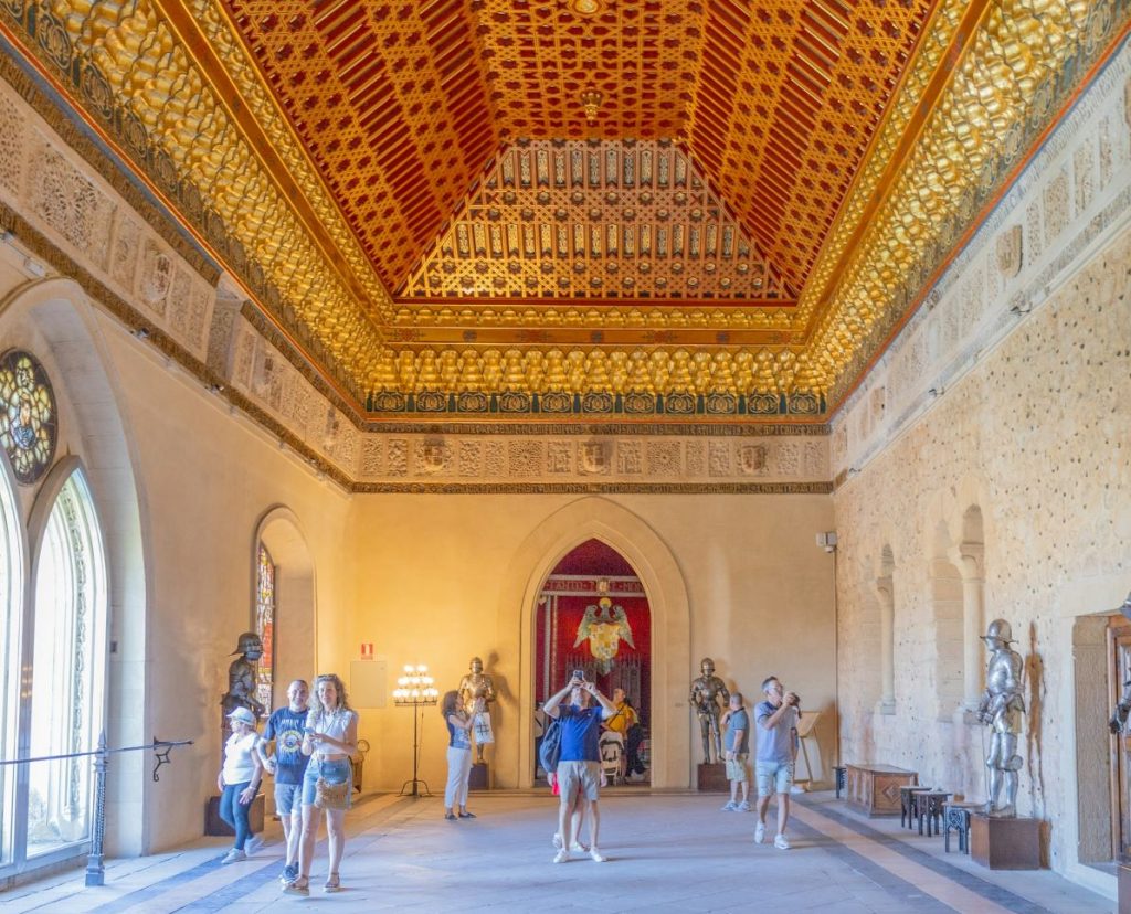 A tour group walks through Alcazar Segovia’s grand hall, featuring golden geometric patterns, statues, and sunlight streaming through arched windows.
