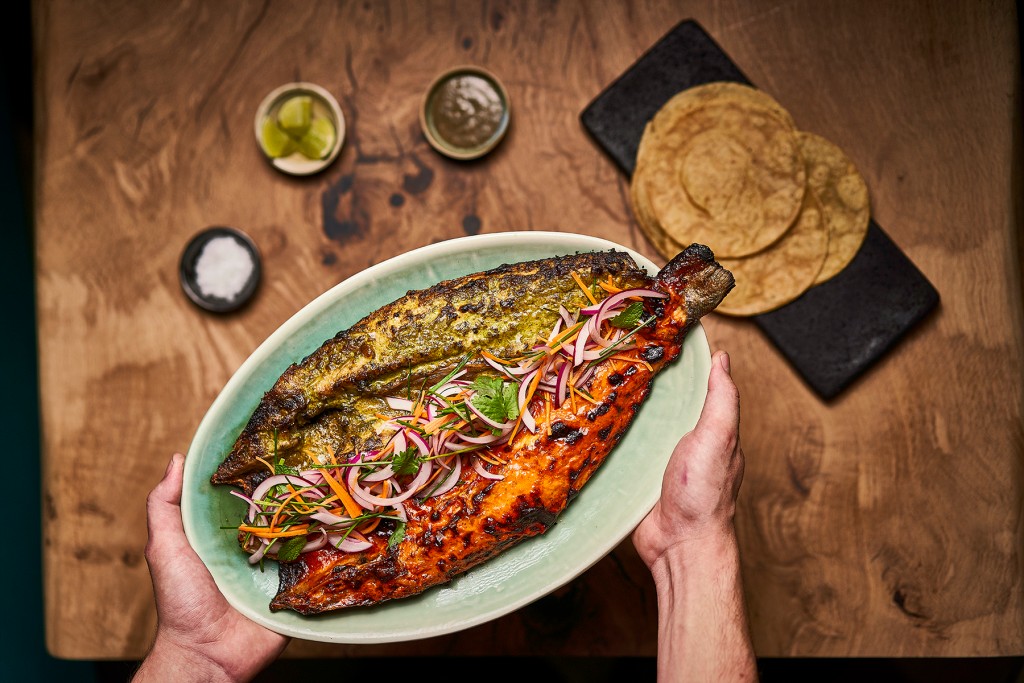 A person presents a whole grilled fish with colorful sauces, onions, and herbs; tortillas, limes, salsa, and salt are nearby.