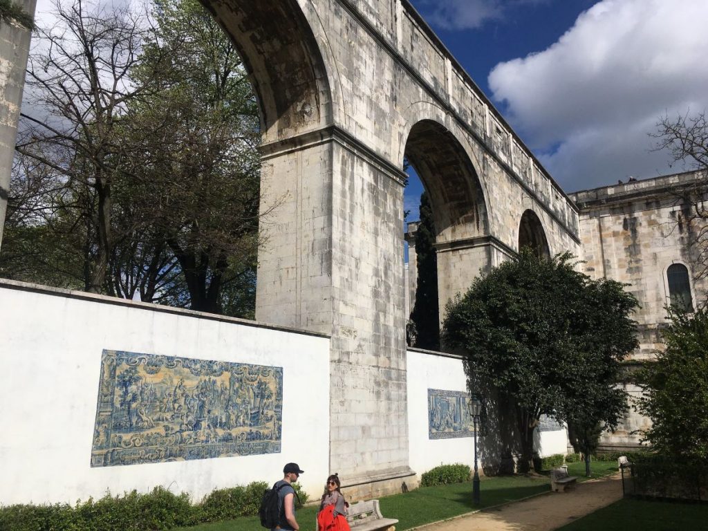A historic stone aqueduct with large arches towers above tiled murals in Lisbon; two people walk beneath amidst trees and clouds.