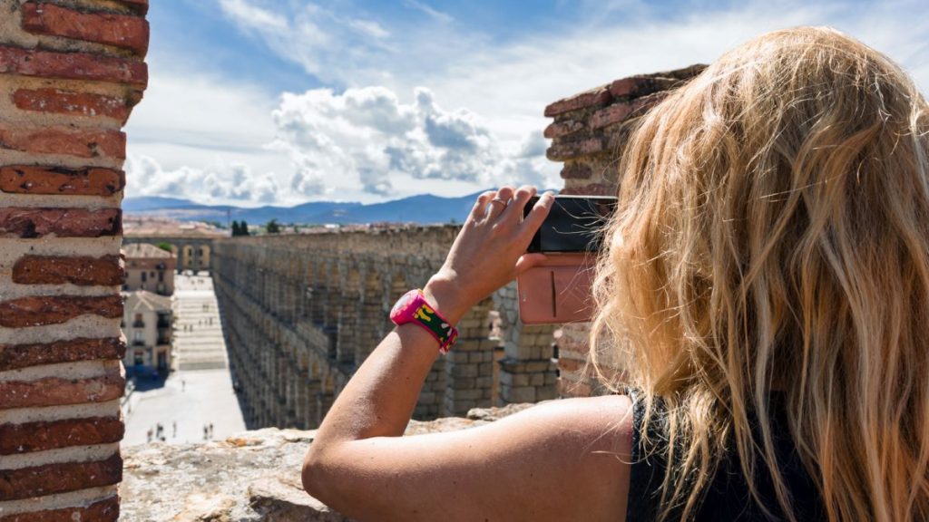 A blonde woman photographs the ancient aqueduct in Segovia, Spain, with the bustling Plaza del Azoguejo and Alcazar nearby.