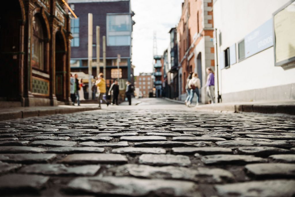 People walk along busy sidewalks beside a cobblestone street, with uneven stones clearly visible in the foreground.