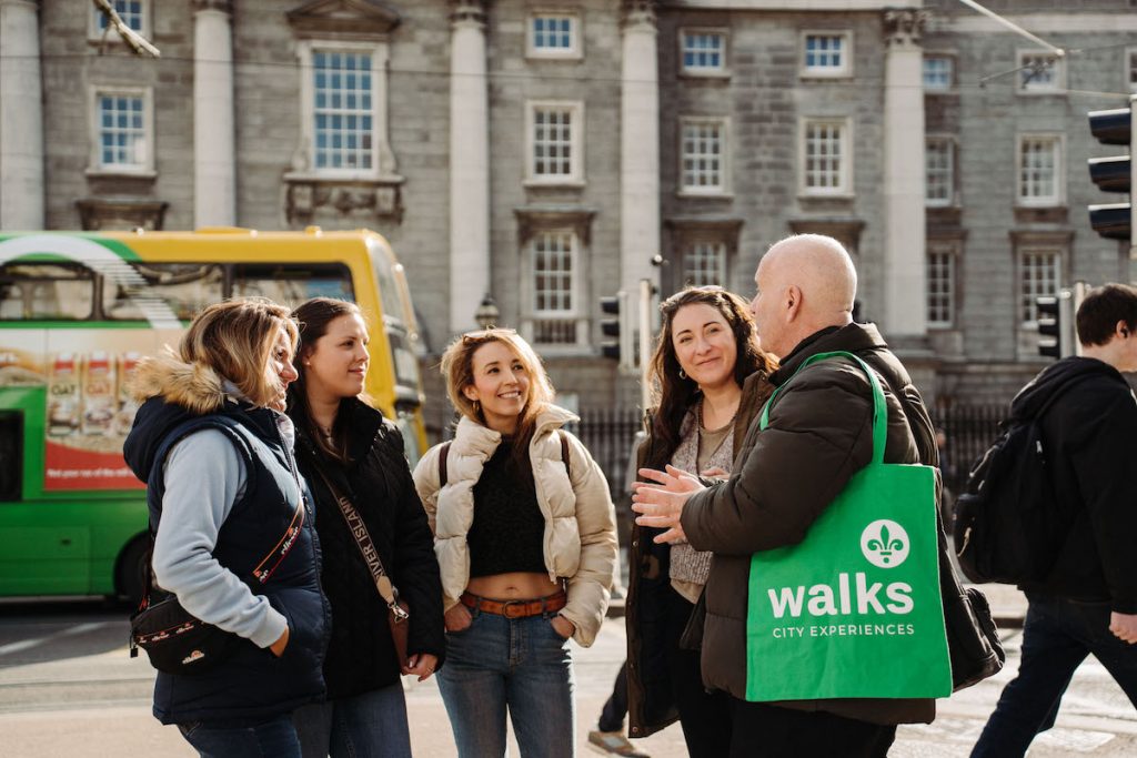 A tour guide with a Walks City Experiences tote engages four smiling women near a green bus and historic stone building.