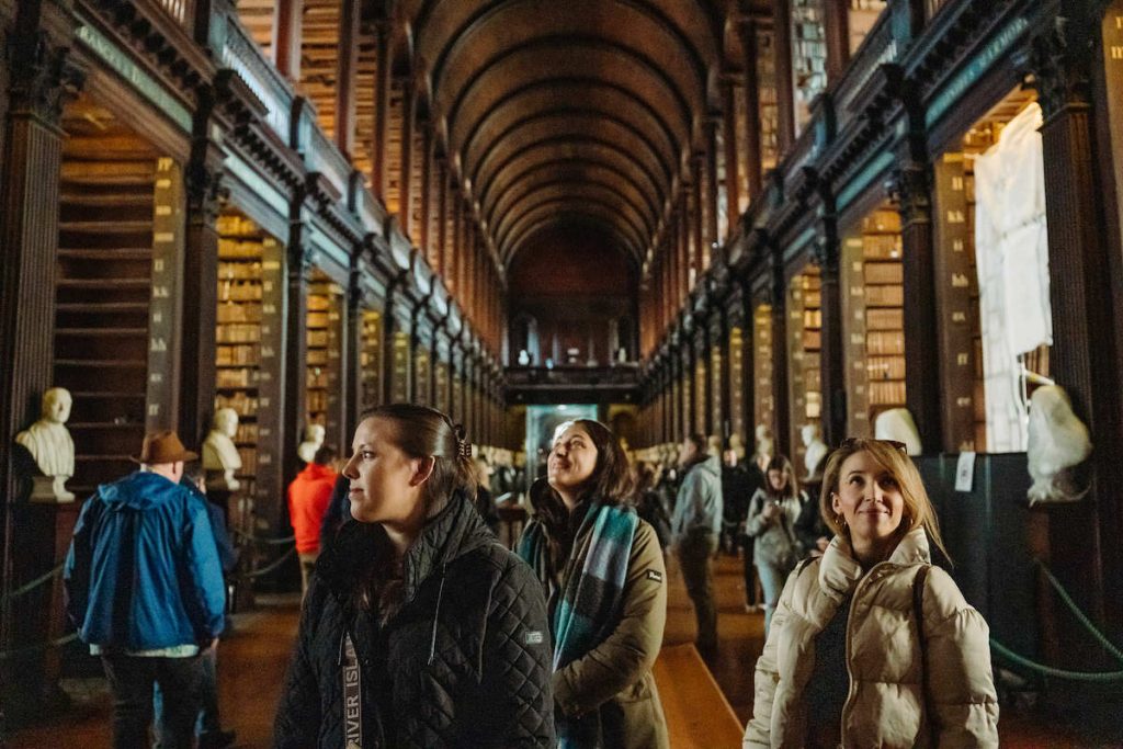 Three women admire tall bookshelves and marble busts in Dublin’s Trinity College Library, known for its historic Long Room.