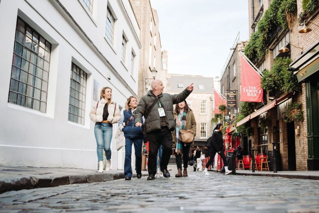 Several travelers follow a tour guide along a cobblestone street in Dublin, passing pubs with red signs and buildings with large windows.