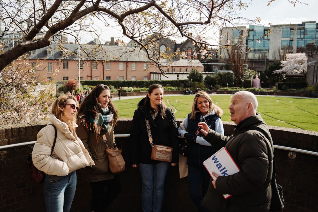 A tour guide talks to four smiling women in a sunny garden near historic buildings and trees, everyone appearing engaged and cheerful.