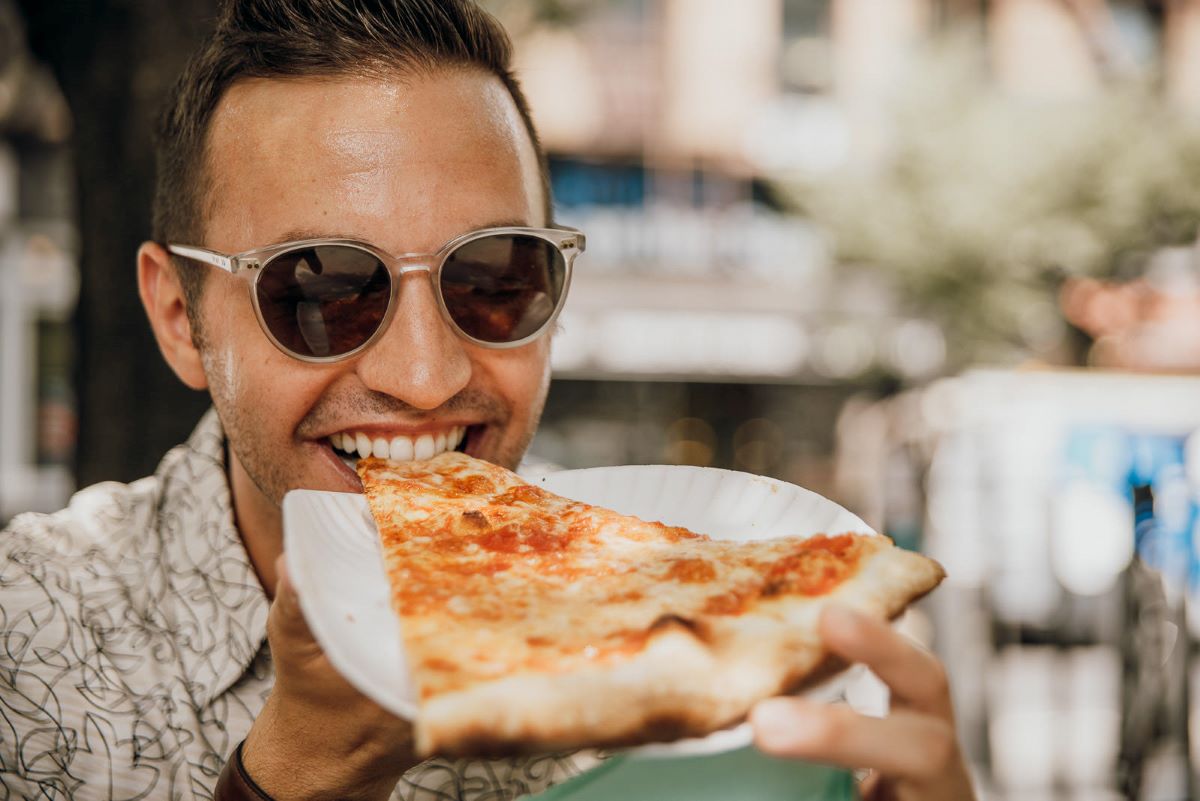 A smiling person wearing sunglasses takes a bite of pizza from a white plate while sitting outdoors at an Astoria restaurant.