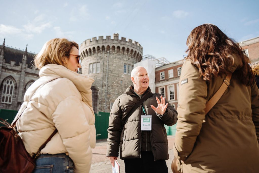 A tour guide explains the history of a stone building with a round tower to two women on a sunny winter day.