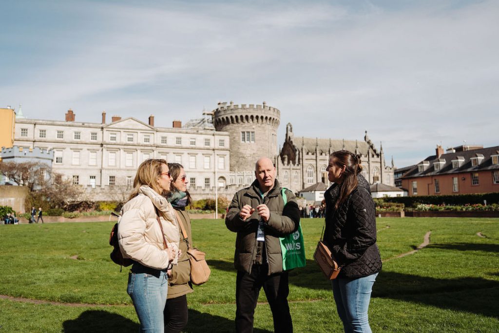 A tour guide explains Dublin Castle’s history to three women on a grassy lawn, surrounded by stone buildings under clear skies.