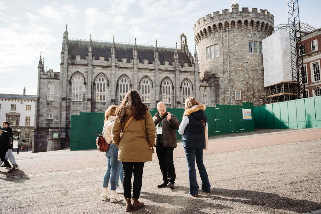 A tour guide addresses four visitors outside Dublin Castle’s stone tower and chapel in Dublin, Ireland, on a sunny day.