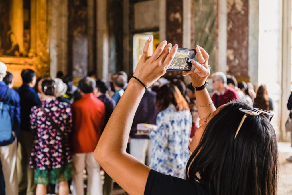 A woman takes a photo with her phone in the crowded Hall of Mirrors at Versailles as sunlight fills the ornate space.