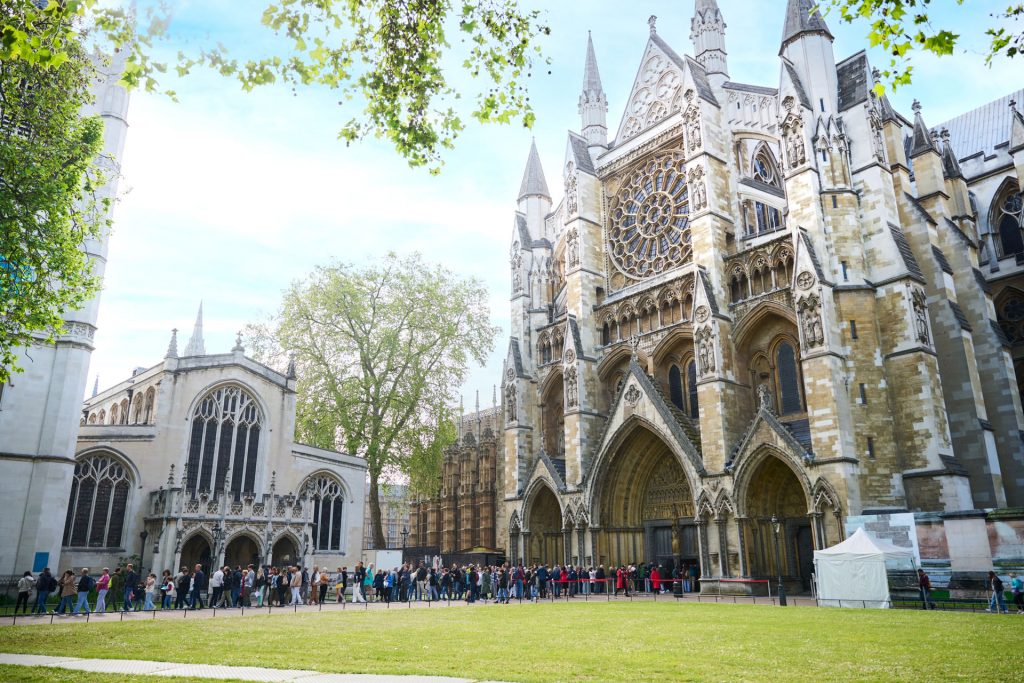 A crowd of people waits outside Westminster Abbey in London, admiring its ornate Gothic architecture and tall spires on a sunny day.