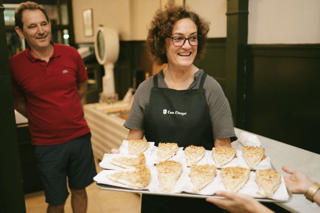 A woman wearing an apron presents a tray of cake slices, while a man in red smiles beside her in a sunny bakery.