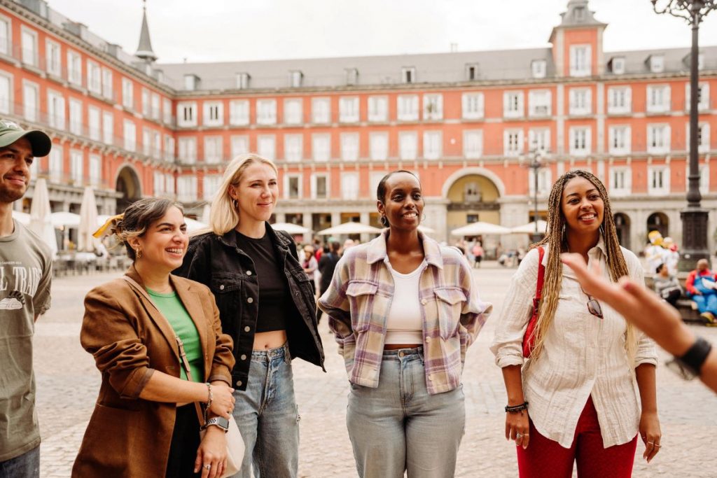 Five people stand in Plaza Mayor, Madrid, smiling and talking together with the plaza’s red and white facade behind them.