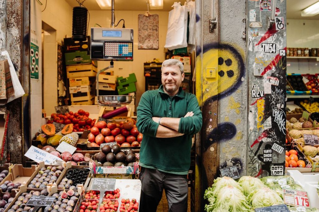 A bearded man in a green sweater smiles with arms crossed at Mercado de San Fernando in Madrid, surrounded by fresh produce.