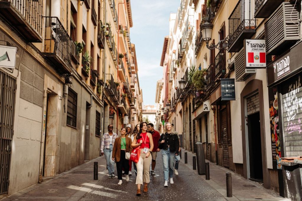 Several people walk along Calle de las Huertas in Madrid, chatting and smiling among colorful buildings with green balconies under sunny skies.
