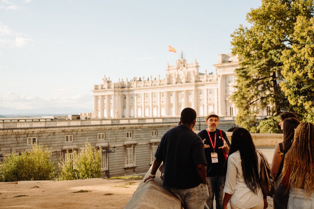 A tour guide speaks to a group outside the Royal Palace of Madrid, with the Spanish flag visible above the palace entrance.