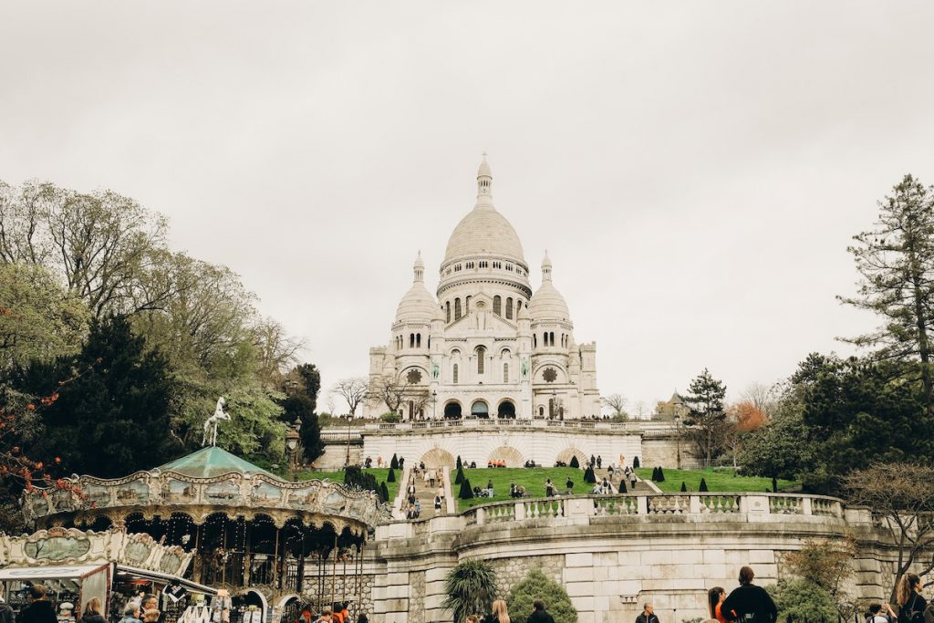 Sacré-Cœur Basilica rises on Montmartre hill in Paris, with visitors relaxing on the lawns and a vintage carousel nearby.
