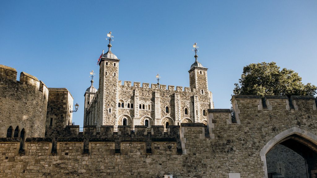 The Tower of London, located in central London, displays its stone walls and turrets beneath a clear sky beside leafy trees.