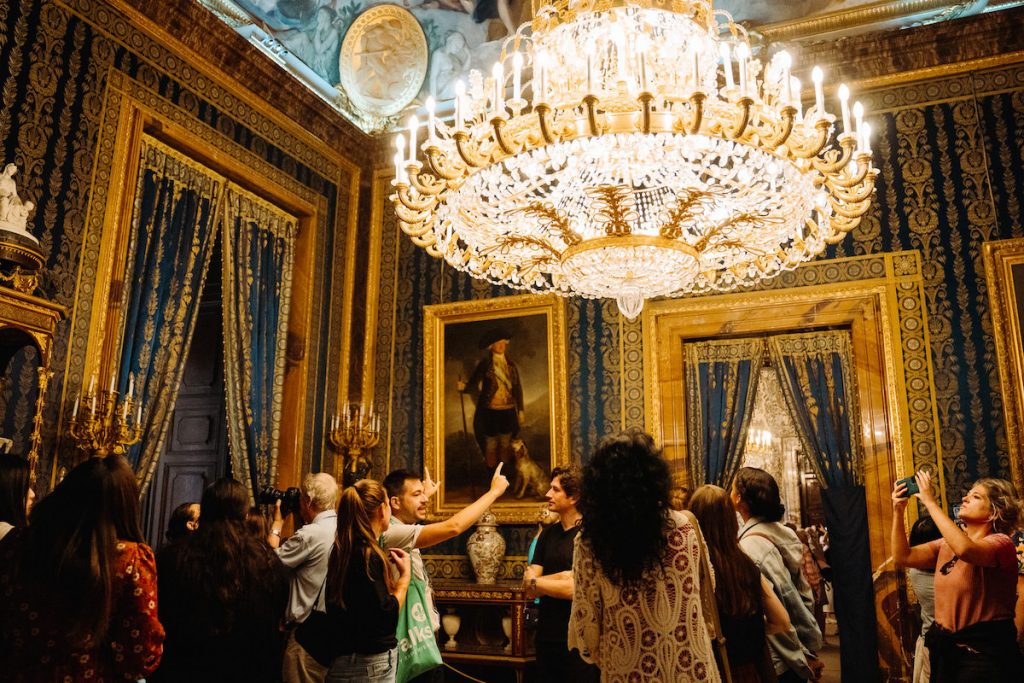 Tour guide explaining a painting in Palacio Real Madrid, where a massive chandelier hands from the ceiling with warm light.