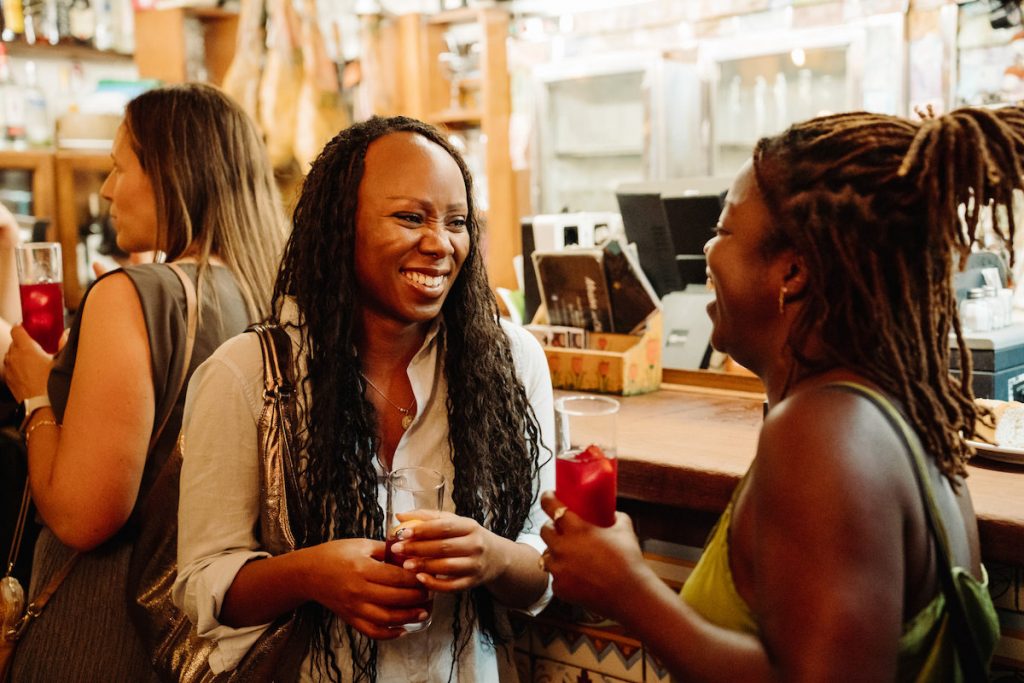 Two women laugh together while holding cocktails at a bar in Madrid, with another guest nearby and vibrant bar decor behind them.
