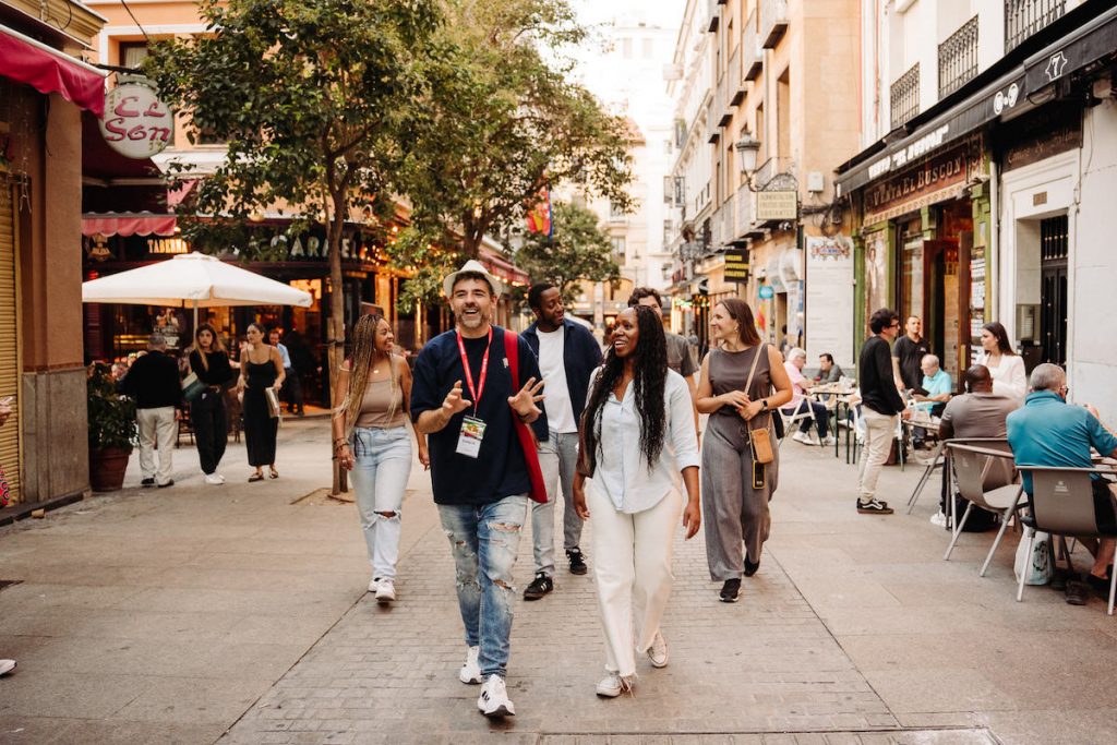 People walk around Madrid's center passing shops and cafes as others relax at outdoor tables under glowing lights.