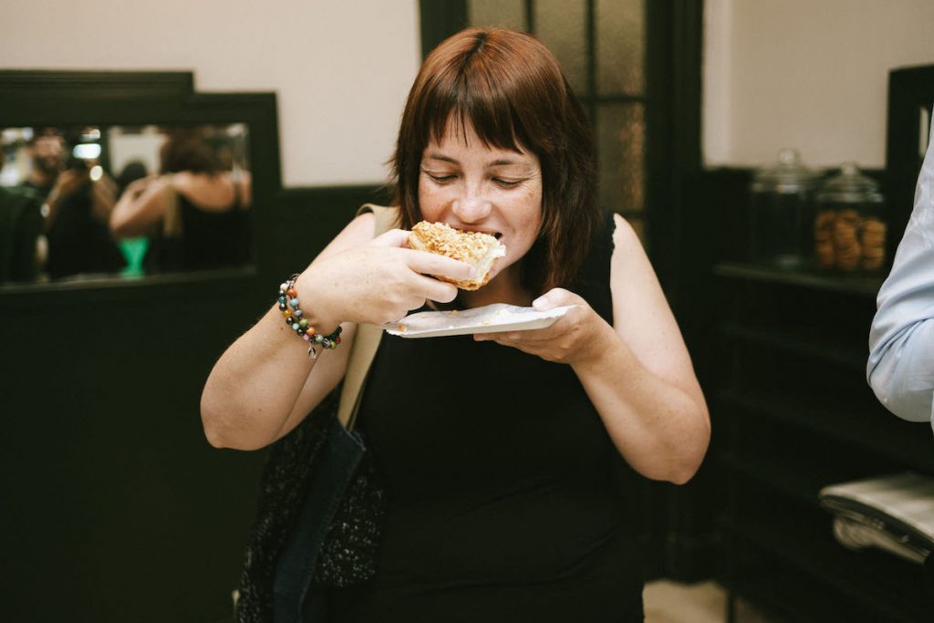 A woman with brown hair and a beaded bracelet eats a sandwich indoors near shelves and a black door in San Sebastián.