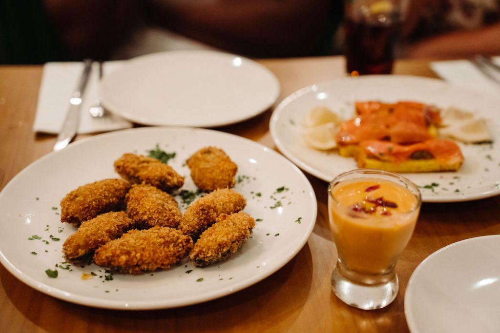 At a typical Spanish tapas bar, croquettes with herbs, smoked salmon on bread, and orange sauce sit on a wooden table.