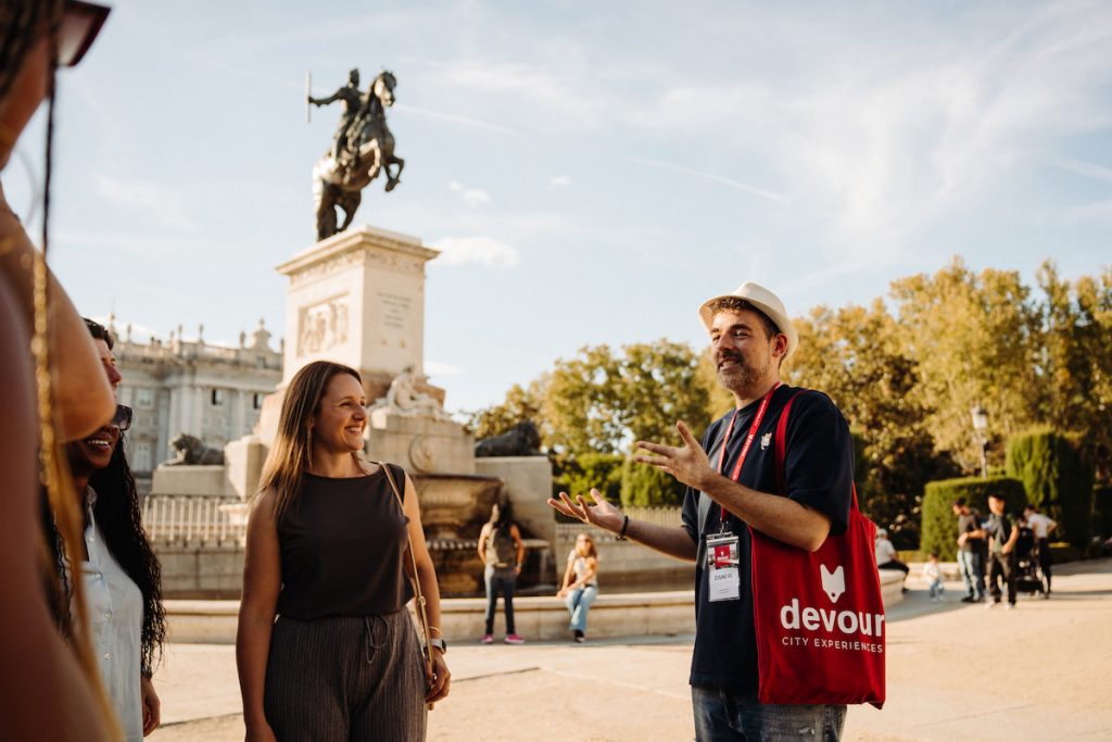 A group of tourists listen to a guide talk, with Plaza de Oriente in the background