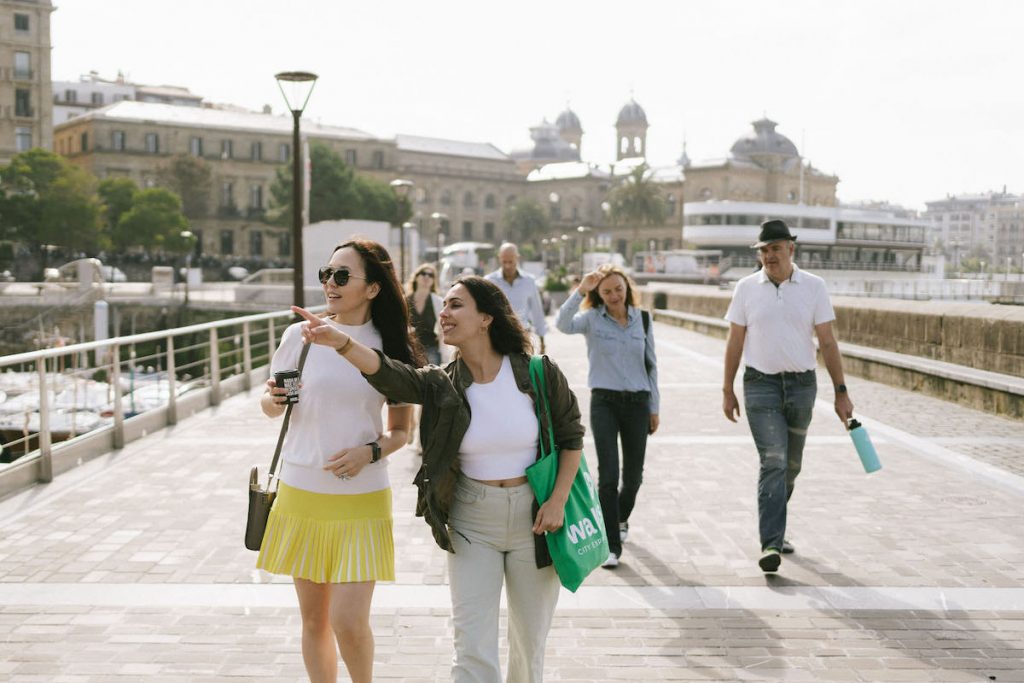Two young women smile and walk along a sunlit riverside promenade; one points ahead. Three others follow, historic buildings behind.
