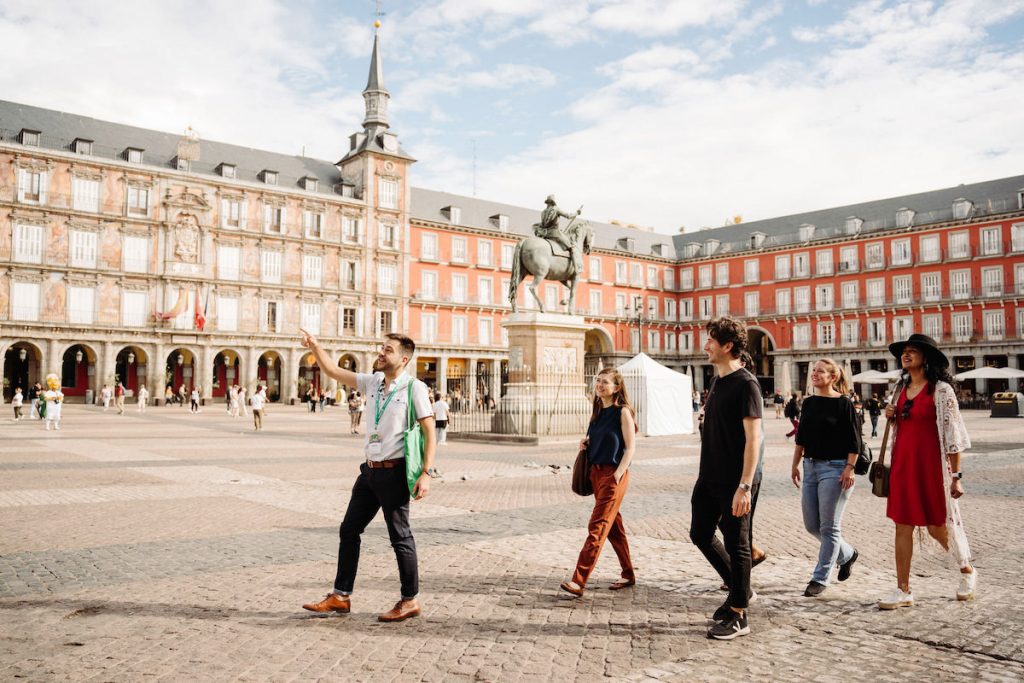 A tour guide leads five people through Plaza Mayor, Madrid, near the equestrian statue and red-and-white historic arcaded buildings.