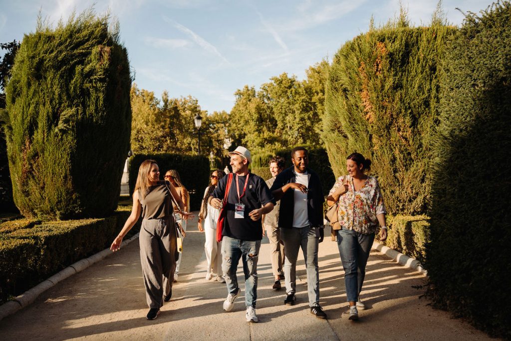Five friends walk and laugh together as they walk in the park bordered by tall, neatly trimmed green hedges.