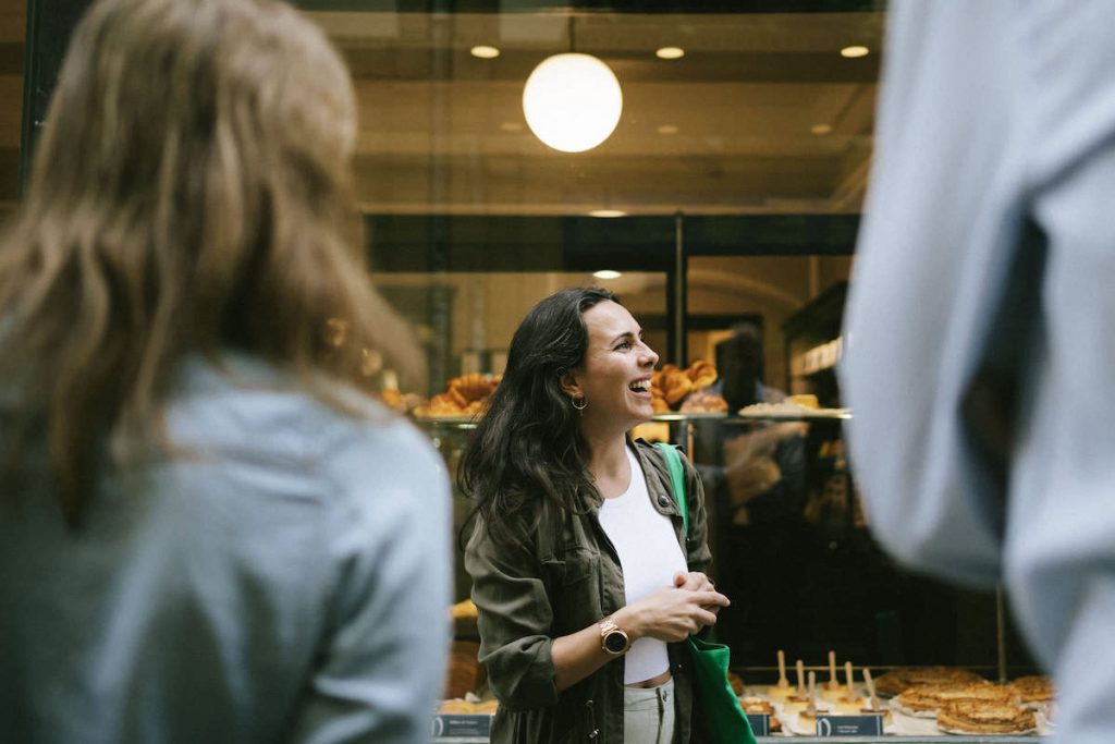 A woman smiles and chats outside a bakery in San Sebastian Old Town, with pastries behind glass and two blurred bystanders.