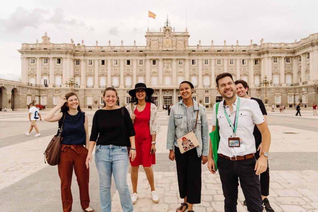 A group of five tourists in front of the Madrid Royal Palace, standing behind their tour guide.