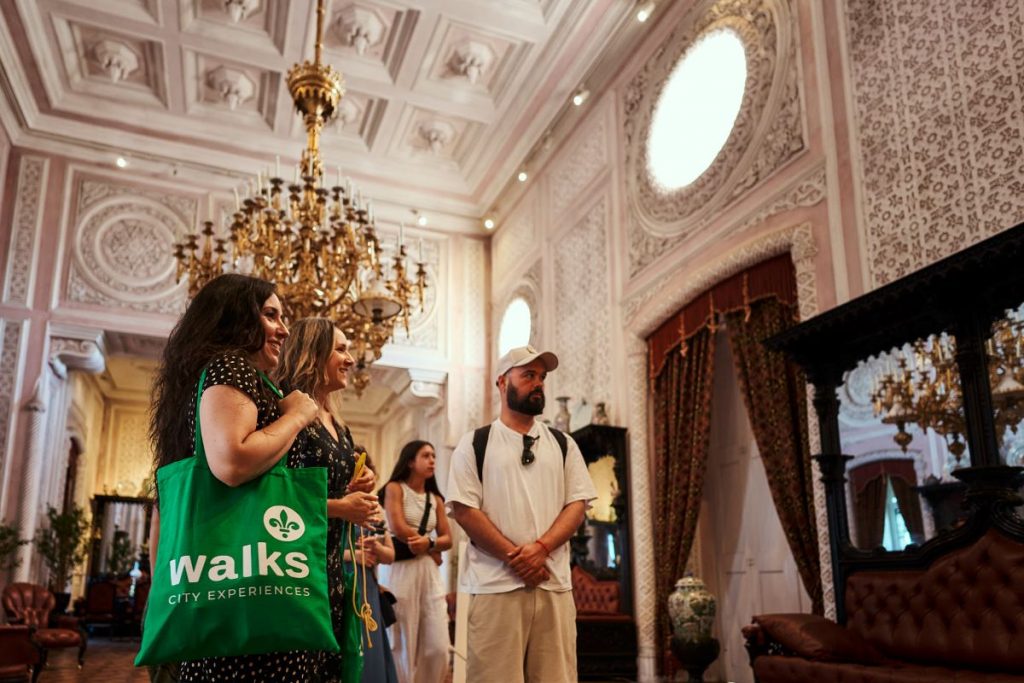 A tour guide explains the history of Pena Palaces lavish, chandelier-lit hall while a woman with a green tote bag listens intently.