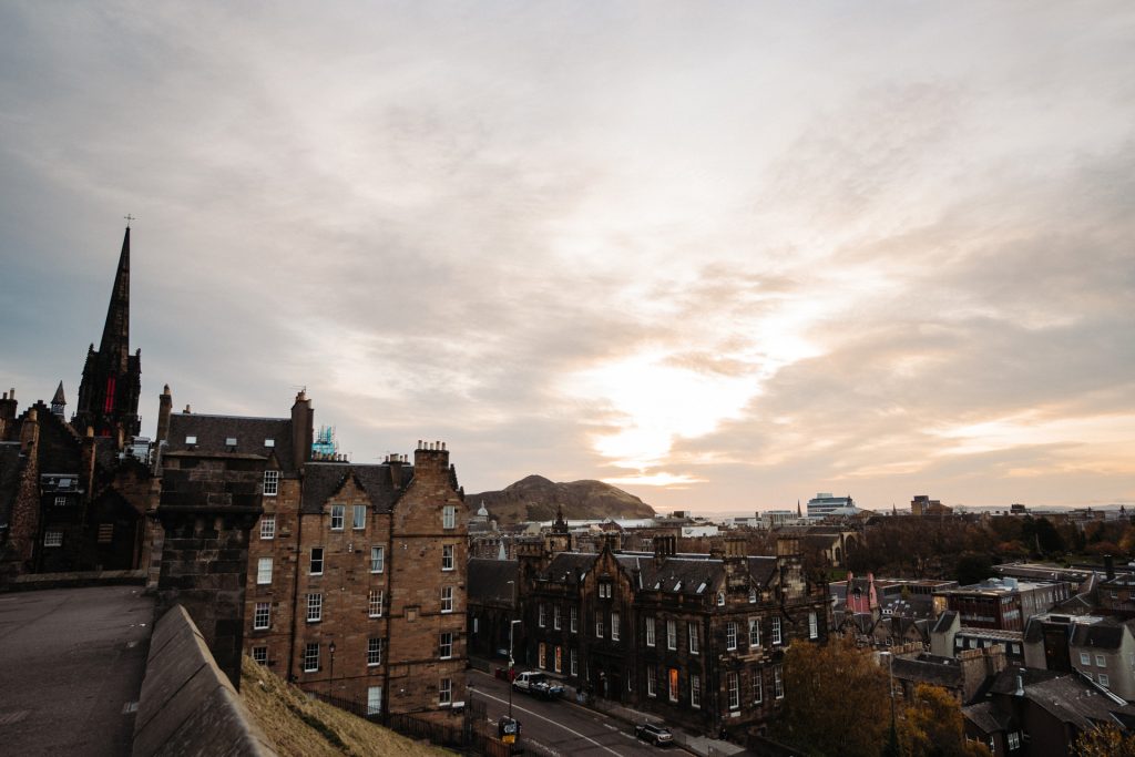 A stunning sunset over Edinburgh, Scotland, reveals historic stone architecture and a prominent church spire against silhouetted hills.