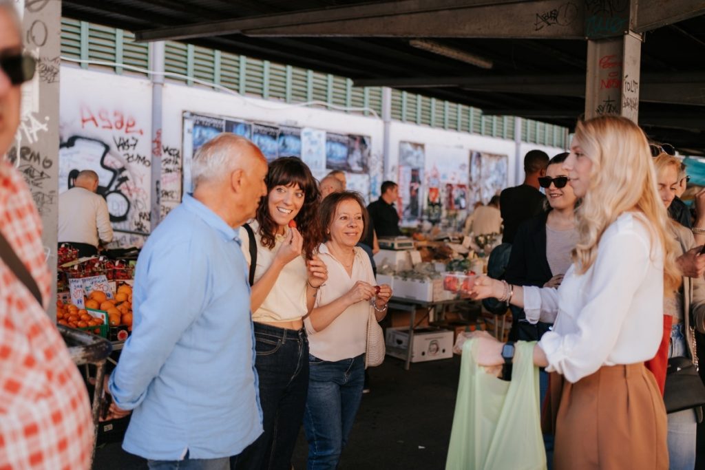 Several people smile and chat near fruit stands at SantAmbrogio Market in Florence, as one woman offers fruit samples.