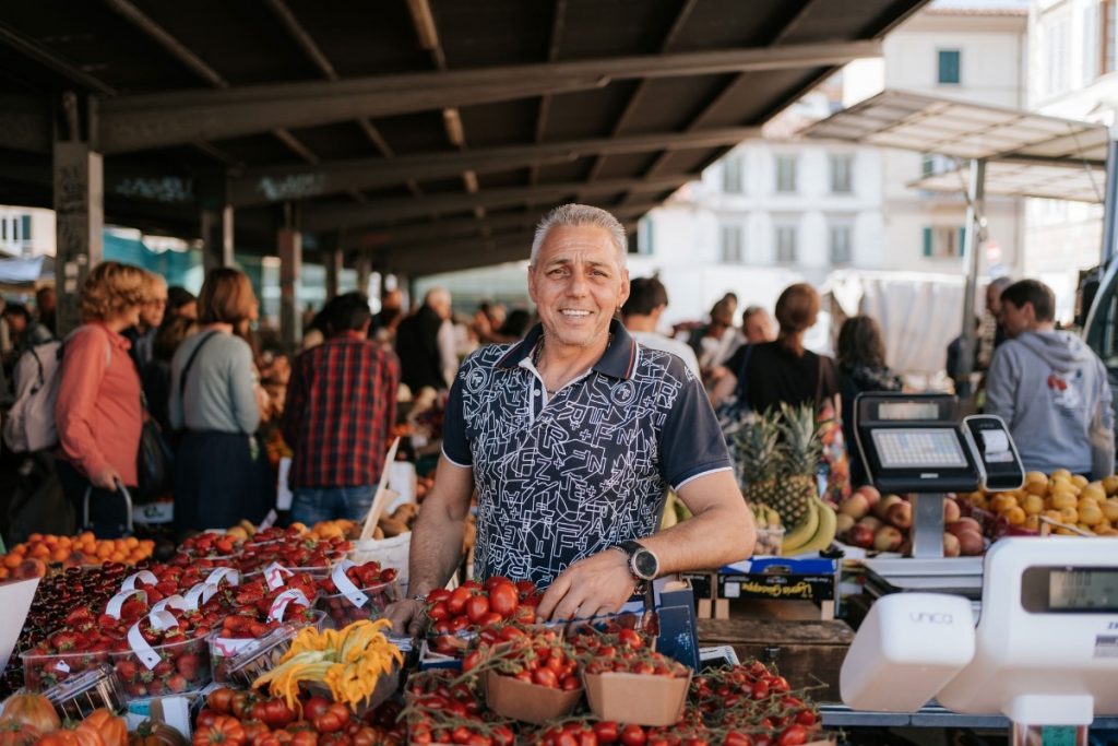 A smiling vendor arranges colorful fruits and vegetables at SantAmbrogio Market in Florence, surrounded by shoppers and busy market stalls.