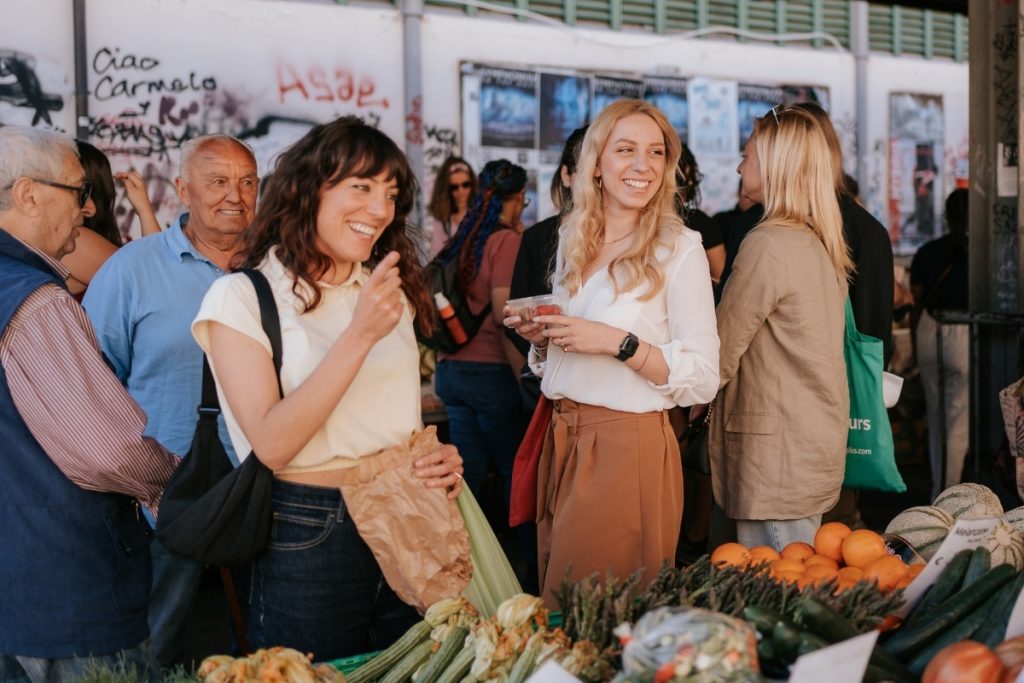 Two women laugh together at SantAmbrogio Market in Florence, examining colorful vegetables amid busy shoppers and graffiti-marked walls.