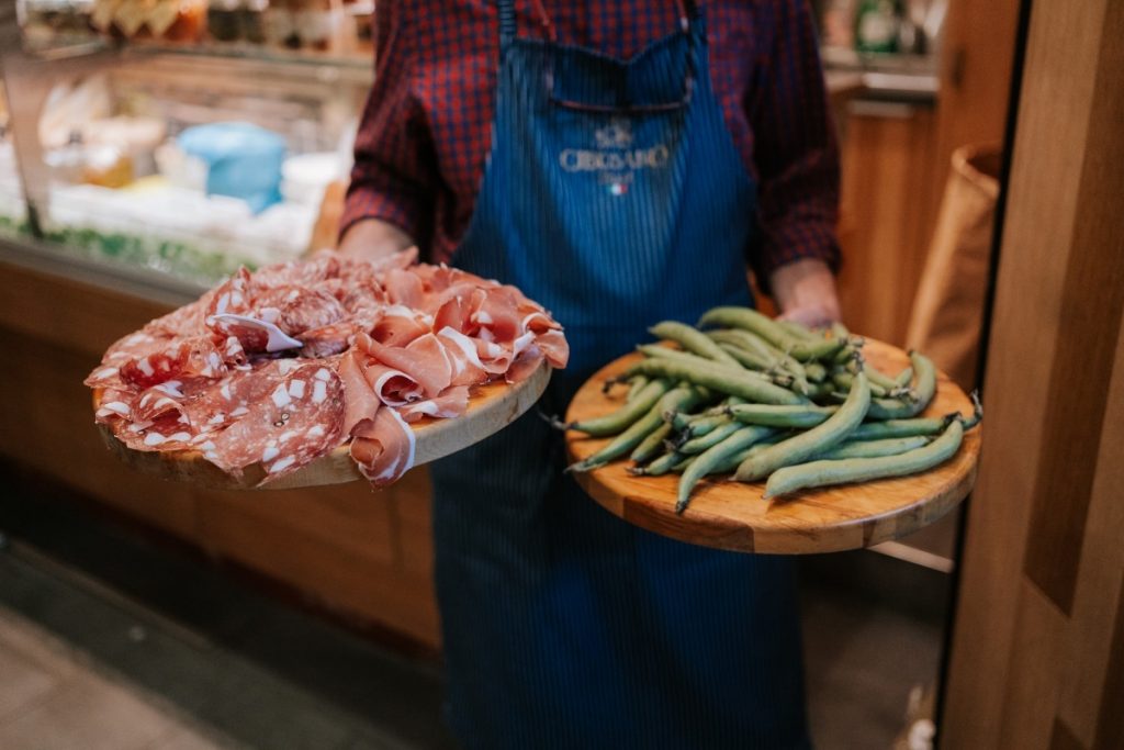 At Florences SantAmbrogio market, a vendor in a blue apron presents cured meats and fresh green beans on wooden platters.