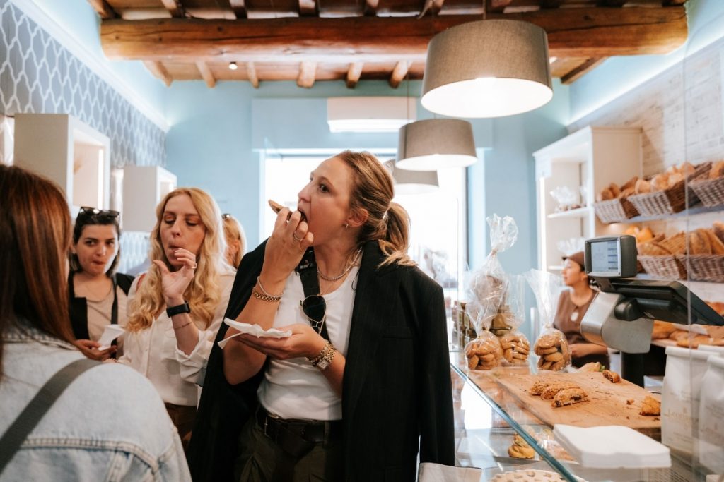 Four women taste pastries inside Pasticceria Giovanni Galli near Florence’s Sant’Ambrogio market; one woman applies lipstick, surrounded by fresh baked goods.