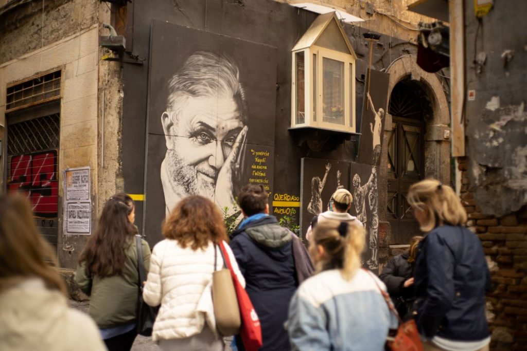Tourists walk down Via dei Tribunali in Naples, passing a large mural of an older man covering one eye.