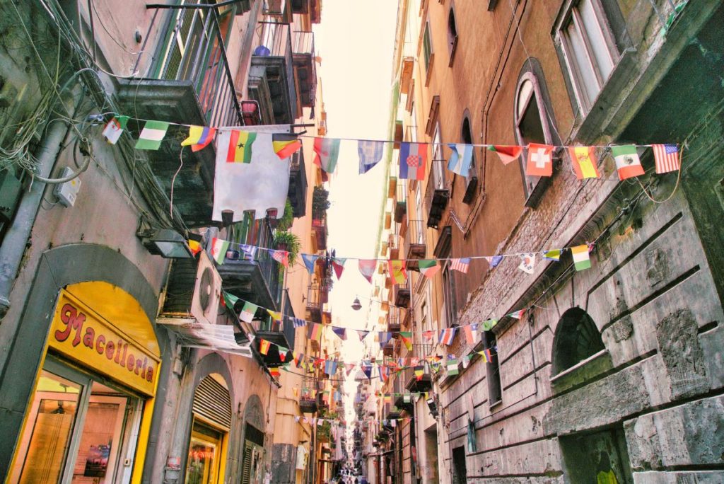 A lively street in Naples’ Quartieri Spagnoli, with laundry strung overhead and Nacotteria’s glowing storefront inviting evening shoppers.