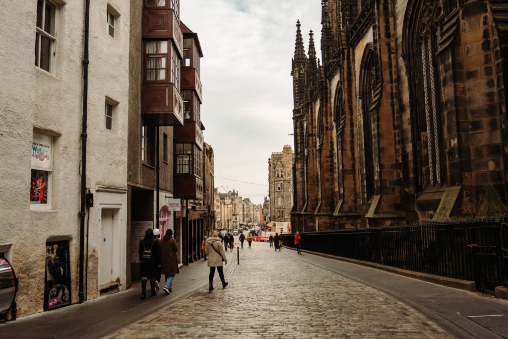 Several people stroll down Edinburgh’s Royal Mile, passing historic stone buildings and the towering St Giles’ Cathedral under gray clouds.