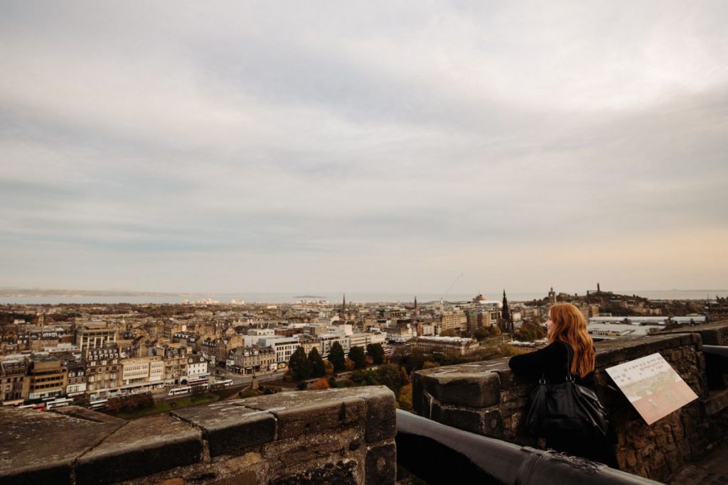 A person with long reddish hair stands at Edinburgh Castle’s stone railing, looking over the city, distant hills, and Firth of Forth.