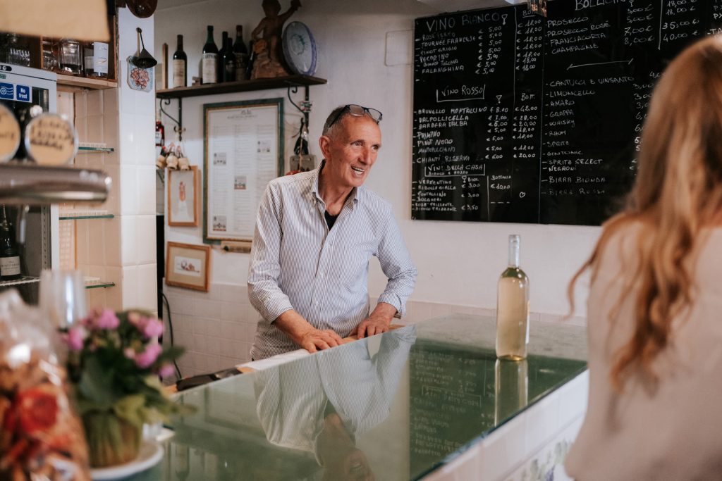 An older man smiles behind a glass counter at Enoteca De Giusti in Florence near SantAmbrogio Market, with wine displayed.