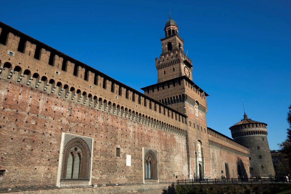 Sforza Castle in Milan, Italy features high brick walls and a large central tower beneath a clear blue sky.