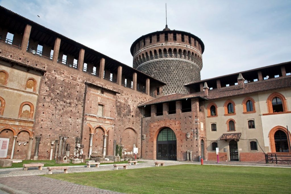 The courtyard of Sforza Castle in Milan features arched windows, grassy lawns, stone benches, and a prominent round tower.