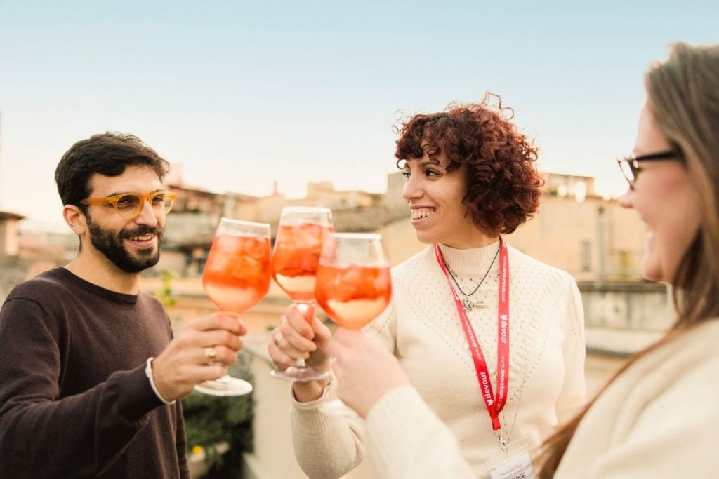 Three friends toast Aperol Spritz at Piazza del Duomo, Milan, with the cathedral and cityscape visible under a sunny sky.