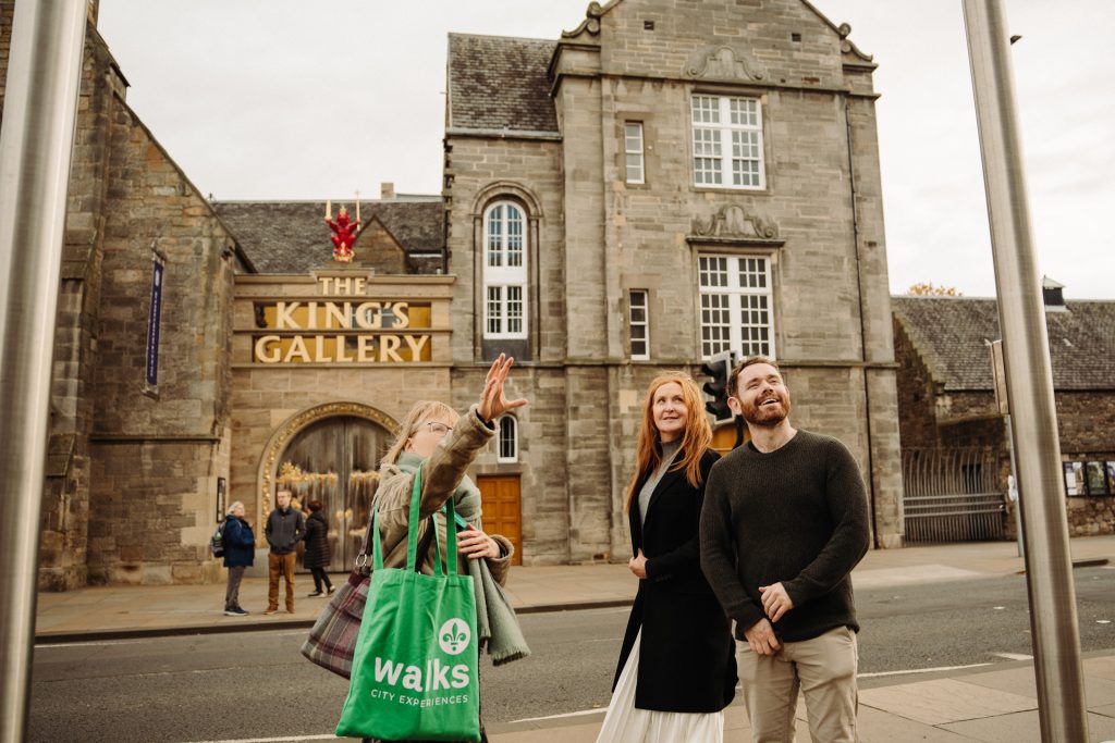 Three people smile outside The King’s Gallery on the Royal Mile in Edinburgh; a woman points up, holding a green Walks tote.