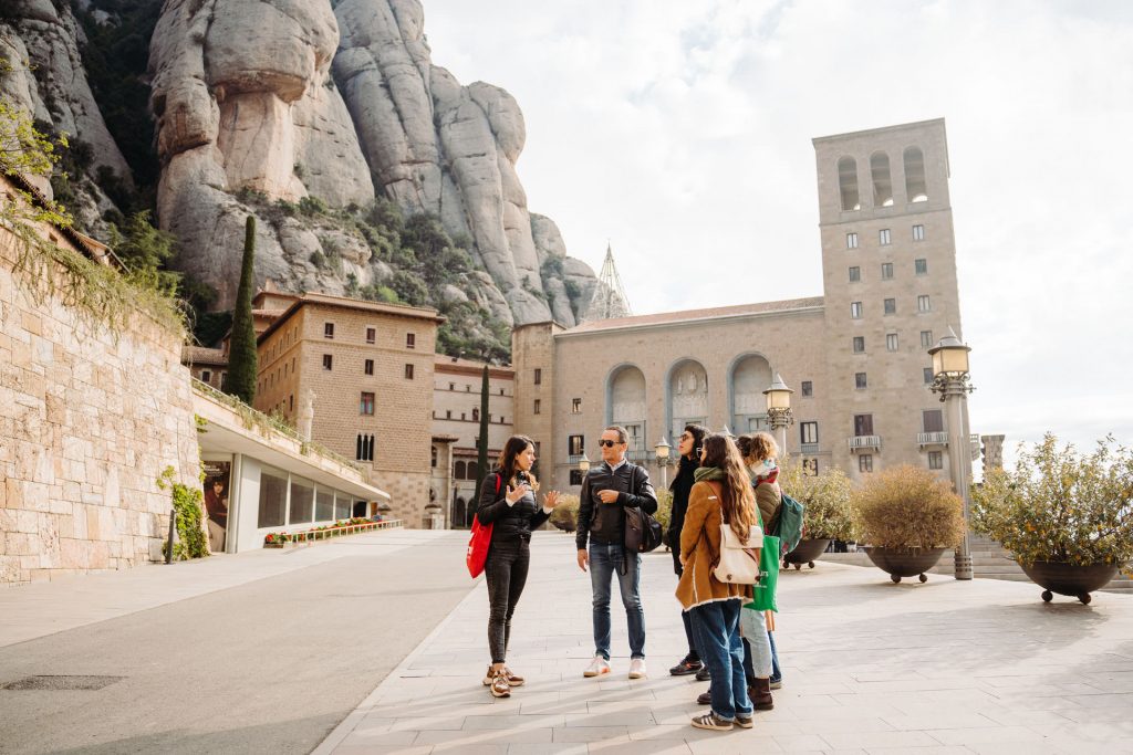 A tour guide speaks to casually dressed visitors in Montserrat’s stone courtyard, with rugged mountains and partly cloudy skies behind them.